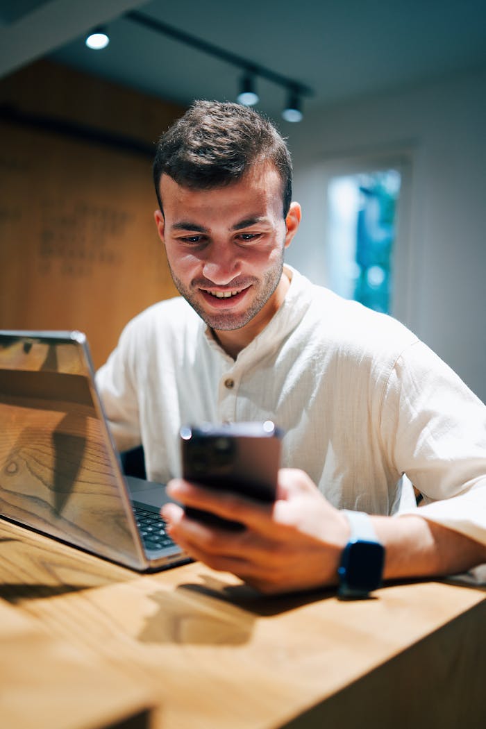 Young man smiling while working on laptop and phone at desk indoors, showcasing modern digital lifestyle.